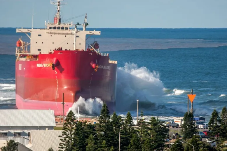 Beach area in foreground with a large ship towering over it on the left side of the image. To the right, the view stretches across the sea to the horizon. A wave is breaking on the ship.