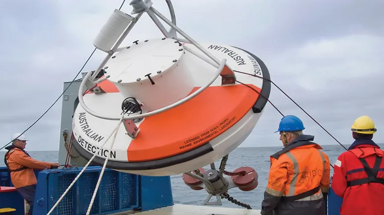 Several people in hi-vis gear and hardhats deploy a deep-ocean tsunami detection buoy from a boat.
