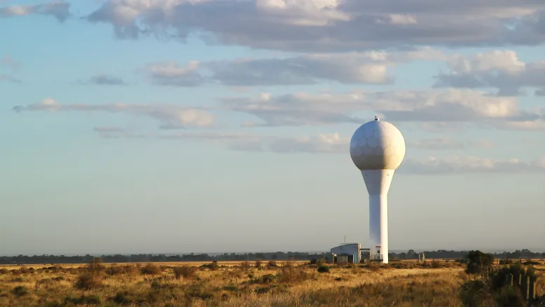White weather radar in grassy rural field environment against a pale blue cloudy sky.