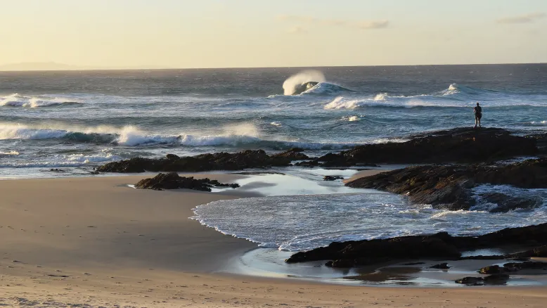 Lone rock fisher stands facing ocean on rocky outcrop close to the beach, with white-peaked waves in background.