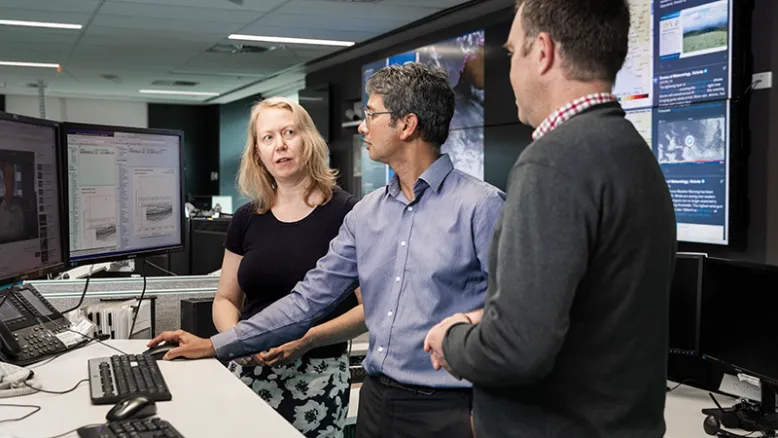 Three Bureau of Meteorology staff members discuss weather images on a computer screen with large screens of the Bureau National Operations Centre behind them.