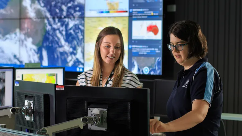 Meteorologist shows computer screen to another staff member. Satellite imagery is visible on screens in background.