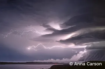 White lightning arcing across a purple sky above the coast. A huge multi-layered storm cloud rises on the right side of the image. Multiple streaks of lightning stretch from the cloud across the sky.