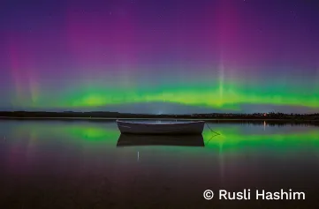 Small boat on still water. Behind the boat, the aurora australis sends colours of green, yellow and purple into the sky. The water reflects the boat and the light, giving the illusion of the aurora stretching across both sky and sea.