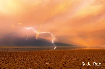 Cracked red dirt on the desert flats of Kimberley. At the left is a dark storm. A lightning bolt crosses a rainbow to strike the ground, arcing from left to the centre of the image. The sun has set on the right, casting orange rays into a pink and gold sky.