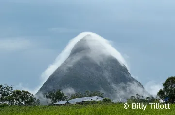 Pyramid-shaped mountain surrounded by dark trees on a green field. Fog has settled like a white blanket of clouds over the mountain, which contrasts with a grey-blue sky. Wind pushes the fog in one direction as it breaks on the rocky slopes.