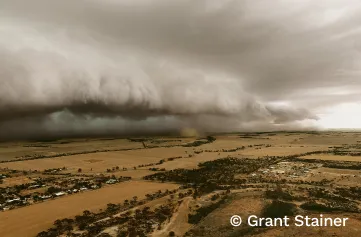 Huge dust and storm cloud approaching houses and trees scattered on a flat farmland plain. Below the band of grey clouds is darkness and shadows, while there is more light where the clouds are yet to reach.