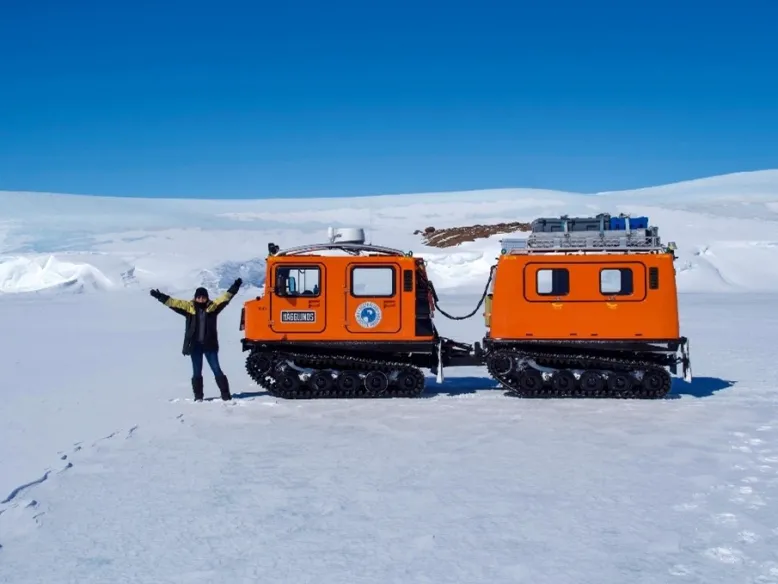 Bureau technical officer stands in front of a bright orange Antarctic vehicle with her arms raised in a snowy landscape.
