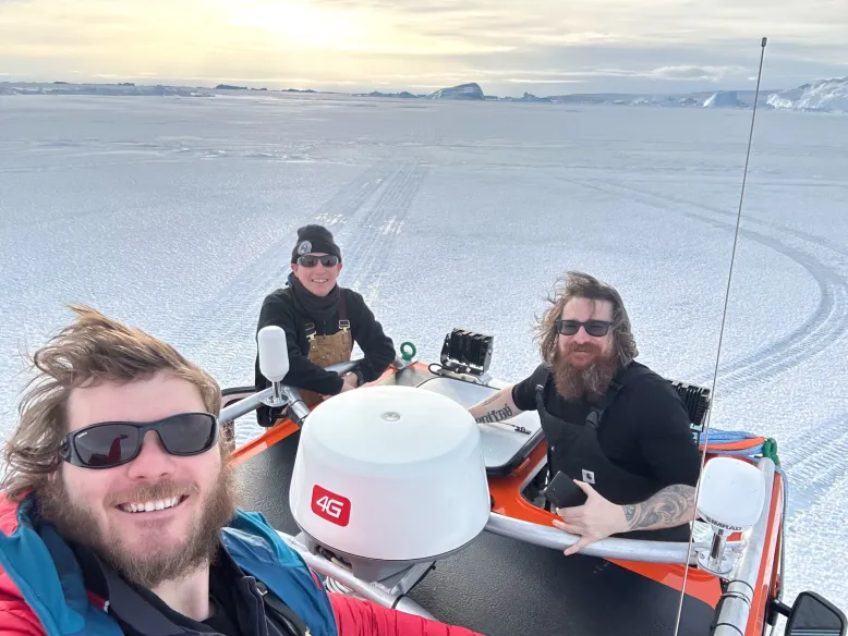 Bureau technical officer sits in a vehicle with two colleagues as they take a windswept selfie on thick sea ice, with a low weak sun in the background.